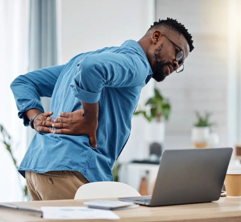 black man with back pain holding both sides of his back looking at a laptop